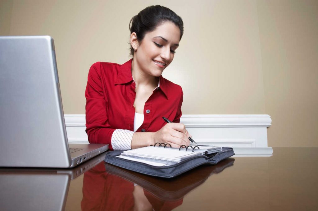 Business woman writing in diary beside laptop in office